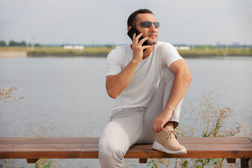 Happy young handsome man sitting on the bench outdoors and using smartphone