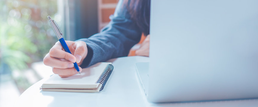 Woman hand is writing on a notepad with a pen and have a laptop computer on the desk in the office.Web banner.