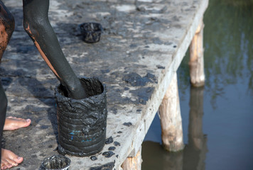 A teenager takes out healing mud from a plastic jar and smears himself with it. The can stands on wooden walkways.