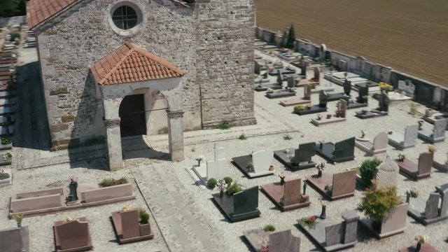Aerial pan parallax shot little town's cemetery with inside the ancient S. Stefano 's Church and bell tower built in XV sec, late-Gothic architecture, zenithal sun light, summer sunny day. Friuli 