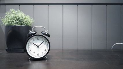 Black color classic alarm clock on the working wood table and decoration with little tree plant for represent work on time or have meeting or have to wake up and nice wallpaper background.