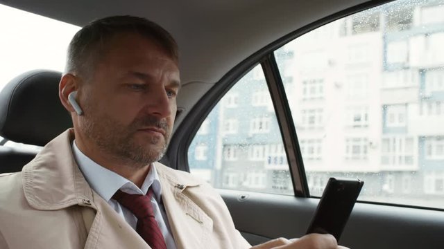 Shaky camera shot of middle-aged businessman in suit and trench coat sitting in back seat of moving car and typing on mobile phone while heading to work