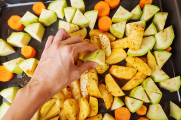 Vegetables - zucchini, potatoes and carrots - lie on a baking sheet sprinkled with spices. Female hand lays vegetables. The concept of cooking vegetarian food, a healthy diet.