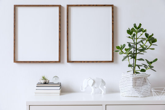 Stylish home interior with two brown wooden mock up photo frames on the white shelf with books, beautiful plants, glassy ball and home accessories. Minimalistic concept of white room decor. 