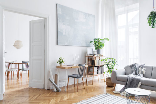 Stylish scandinavian open space with design furniture, plants, bamboo bookstand and wooden desk. Brown wooden parquet. Abstract painting. Modern decor of bright room next to dining room. 