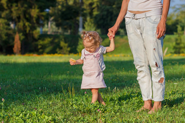 Little cute girl does her first steps with mother support, green grass, summer time, blurred background