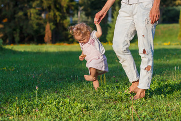 Little cute girl does her first steps with mother support, green grass, summer time, blurred background