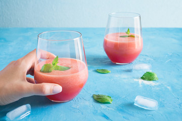 Female hand holds watermelon-melon strawberry smoothie. Summer cocktail with ice and mint on a blue turquoise background with copy space