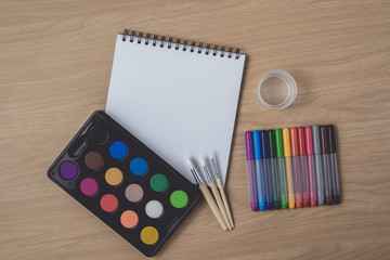 Notepad or notebook with Many colorful pens, paintbrush and Watercolor palette on brown wood table.using for Arts and Education background