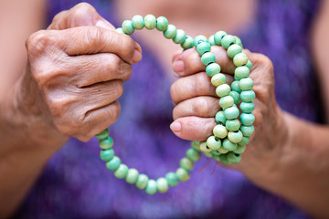 Senior woman's hands praying and holding a turquoise beads rosary in bokeh blue swimming pool...