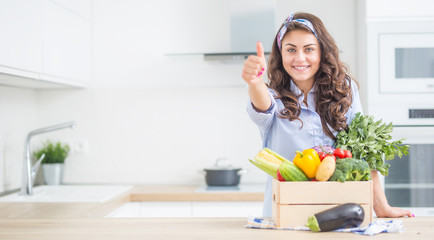 Woman in her kitchen with wooden box full of organic vegetable showing thumbs up