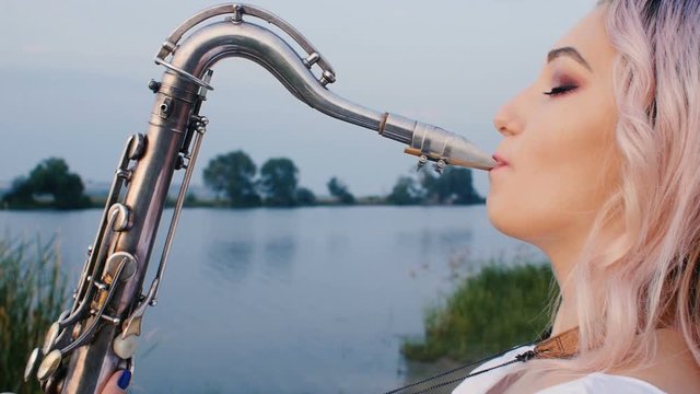 young woman playing on the saxophone near the riverbank at dawn, a beautiful girl in a long dress with a wind musical instrument in nature, a concept of hobby and relaxation, art, inspiration