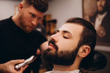 Hair beard and mustache treatment in barber shop. Young, bearded, muscular,  handsome barber making haircut of attractive bearded man in barbershop