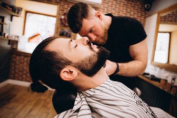 Hair beard and mustache treatment in barber shop. Young, bearded, muscular,  handsome barber making haircut of attractive bearded man in barbershop