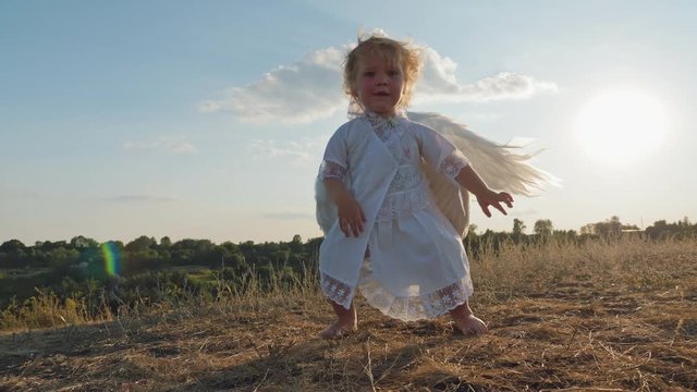 The Child And Mother Like Angels Walk In Nature. A Little Girl Looks At The Camera And Smiles, Against The Background Of A Mom In An Angel Costume, Whirling Around Waving Wings