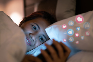 Asian social media influencer woman using smartphone in bed with emoji - Millennial girl browsing internet online at night with emoticon reactions glowing above phone - Web, icons & lifestyle concept
