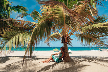 Young woman sit on sand on troical sand pech in the shadow of palm tree. Woman under palm tree at beach