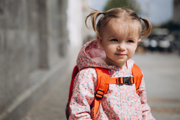 little pretty baby who got lost in the city, a girl in a pink raincoat, with a backpack and a stylish hairstyle sitting next to a large administrative building one by one