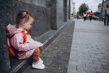 little pretty baby who got lost in the city, a girl in a pink raincoat, with a backpack and a stylish hairstyle sitting next to a large administrative building one by one