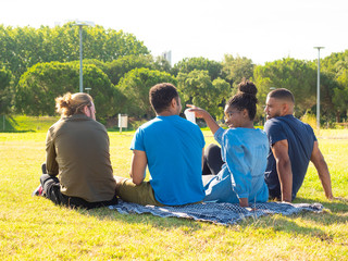 Back view of multiethnic friends gathering in park. Rear view of happy young male and female...