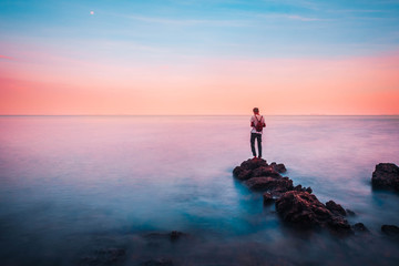 young man stand on a rock in the foreground of him have the sea and sky brightly colored look like...