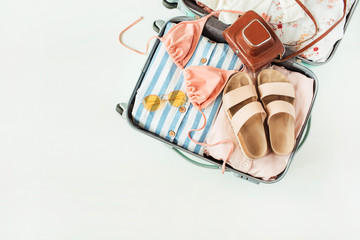 Travel hand luggage with swimsuit bikini, slippers, sunglasses, dress, retro camera on white background. Flat lay, top view summer vacation concept.
