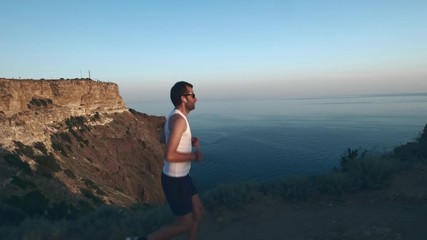 Athletic Handsome Man is Doing Sports with Phone and Headphones, Running along Sea Coast at Sunset while Jogging on Beach in Summer. Uses wireless headphones