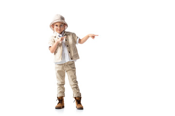 full length view of smiling explorer boy in hat and glasses pointing with fingers isolated on white
