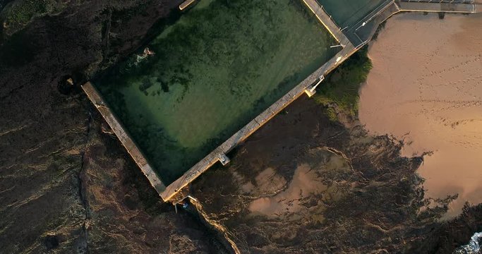 Sports infrastructure rock pool in sandstone plateau on the tip of sandy Mona Vale beach in aerial lifting from the surface to horizon and sky.