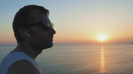 A man runs along the shore, playing sports at sunset. Athlete Listening Music. Athlete Using Wireless Earphones. Sportsman Listening Music In Wireless Headphones. Listening Music On His Smartphone.