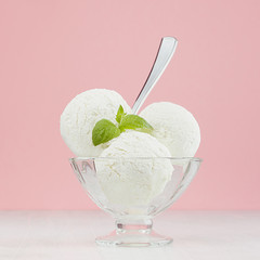Sweet creamy ice cream in elegant transparent glass bowl with green mint, silver spoon on white wood table and pastel pink wall, square, closeup.