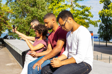 Mix raced people with smartphones enjoying leisure outdoors. Men and women sitting on parapet outdoors, using phones and taking selfie. Gadget addiction concept