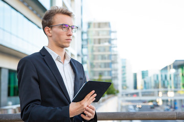 Pensive project leader with tablet thinking over report outside. Young business man standing near office building, leaning on railing, holding tablet and staring into vacancy. Working outdoors concept