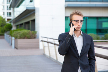 Serious focused office worker calling on phone and getting important news. Young man in glasses and formal jacket standing outside, talking on cell and listening carefully. Important news concept