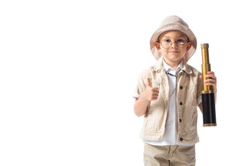 front view of smiling explorer boy in glasses and hat holding spyglass and showing thumb up isolated on white