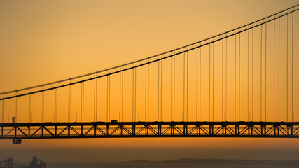 Silhouette of a part of the "Ponte 25 de Abril" (25 de Abril Bridge) with cars against golden background at sunset, Lisbon, Portugal