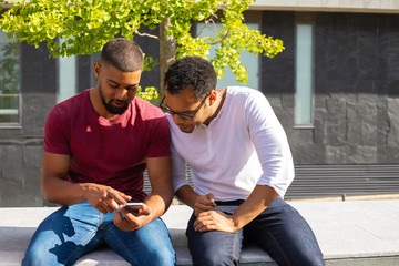 Excited male friends studying mobile app features on phone. Two men in casual sitting on parapet outside, using one smartphone and staring at screen. Mobile app concept