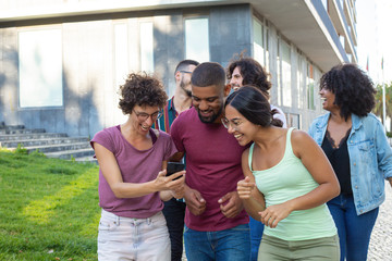 Obraz premium Joyful woman sharing good news with two happy friends and showing smartphone screen. Three laughing people walking behind them outdoors. Good news concept