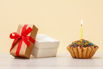 Gift boxes and birthday muffin with burning festive candle on beige background.