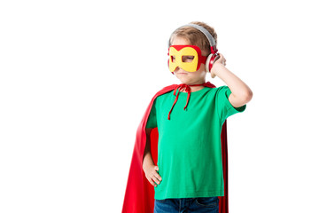 preschooler boy in red hero cloak and mask listening music in headphones and looking away isolated on white