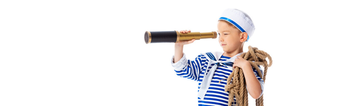 panoramic shot of focused boy in sailor costume holding rope and looking through spyglass isolated on white