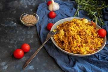 A large plate filled with Uzbek pilaf, next to cherry tomatoes, a dark cloth, fresh pepper in a manually knitted stand. In a transparent dish, spices - zeros and a mixture of peppers. 