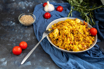 A large plate filled with Uzbek pilaf, next to cherry tomatoes, a dark cloth, fresh pepper in a manually knitted stand. In a transparent dish, spices - zeros and a mixture of peppers. 