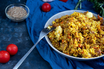 A large plate filled with Uzbek pilaf, next to cherry tomatoes, a dark cloth, fresh pepper in a manually knitted stand. In a transparent dish, spices - zeros and a mixture of peppers. 