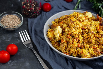 A large plate filled with Uzbek pilaf, next to cherry tomatoes, a dark cloth, fresh pepper in a manually knitted stand. In a transparent dish, spices - zeros and a mixture of peppers. 
