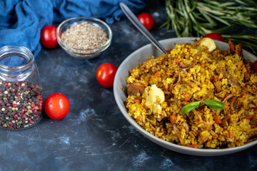 A large plate filled with Uzbek pilaf, next to cherry tomatoes, a dark cloth, fresh pepper in a manually knitted stand. In a transparent dish, spices - zeros and a mixture of peppers. 