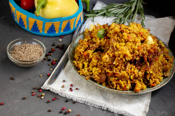 A large plate filled with Uzbek pilaf, next to cherry tomatoes, a dark cloth, fresh pepper in a manually knitted stand. In a transparent dish, spices - zeros and a mixture of peppers. 