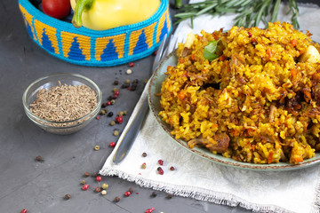 A large plate filled with Uzbek pilaf, next to cherry tomatoes, a dark cloth, fresh pepper in a manually knitted stand. In a transparent dish, spices - zeros and a mixture of peppers. 