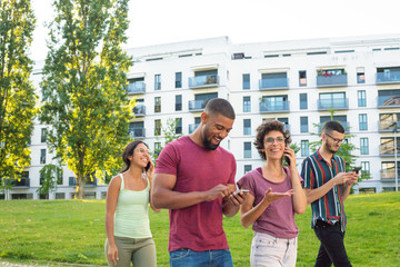 Interracial group of cheerful people using smartphones and talking on cells. Men and women with mobile phones walking in residential area. Communication concept