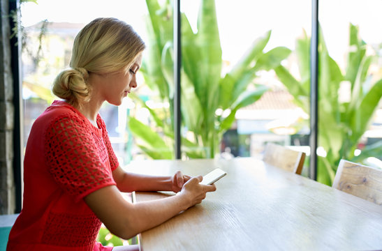 Lifestyle shot of young blonde caucasian natural millennial woman working on cellphone at a modern tropical and bright office with plants